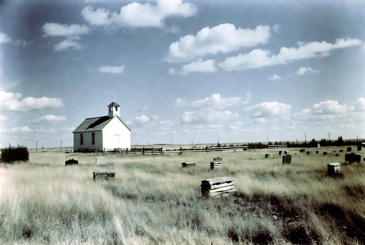 A small white wooden prairie church and a fenced graveyard under a wide sky