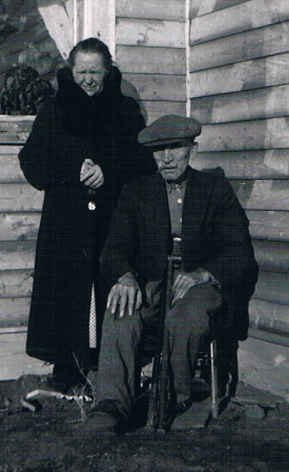 Edward Steinke seated in a flat cap with Augusta standing at his shoulder, beside the farmhouse wall