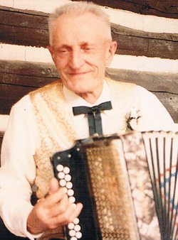 Henry Steinke in a vest and bow tie, smiling over his button accordion against a log wall